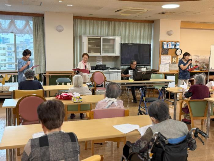 施設内でボランティアが演奏している。参加者の座席間は空いており、マスクを着けている。