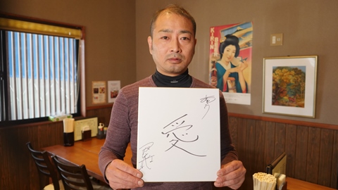 「愛」と書かれた紙を持った塚本さんの画像